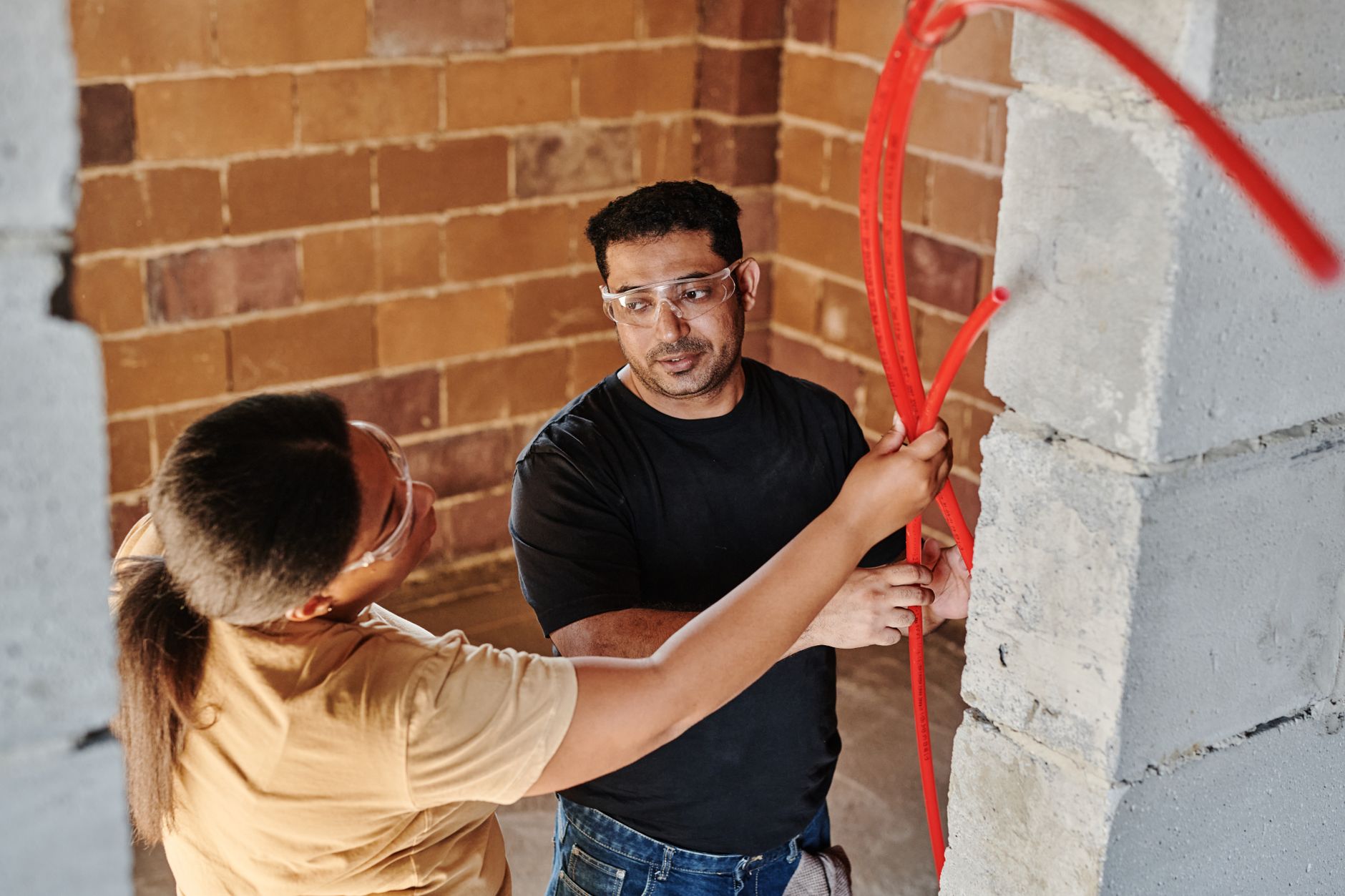 Two workers fitting red tubing on a brick wall at a construction site